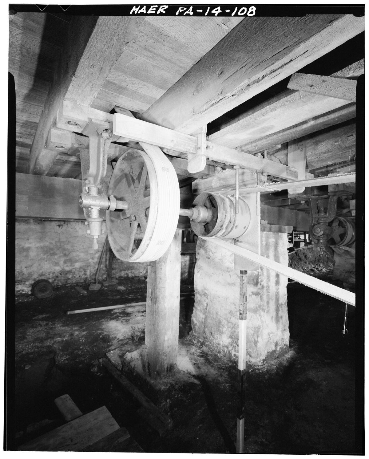 A horizontal drive shaft and a large belt pulley in a factory basement, with a belt running up under the wooden ceiling beams.